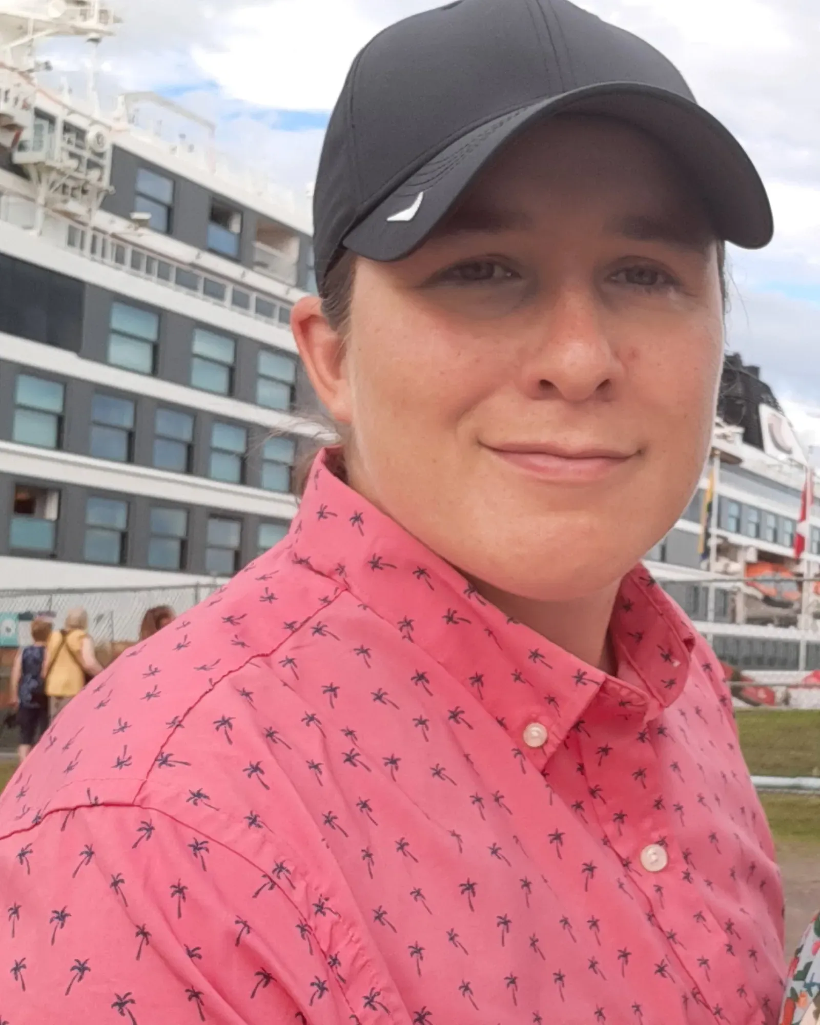 Jamie Scrimger outdoors in a ball cap and patterned shirt, a cruise ship docked at the waterfront behind her.
